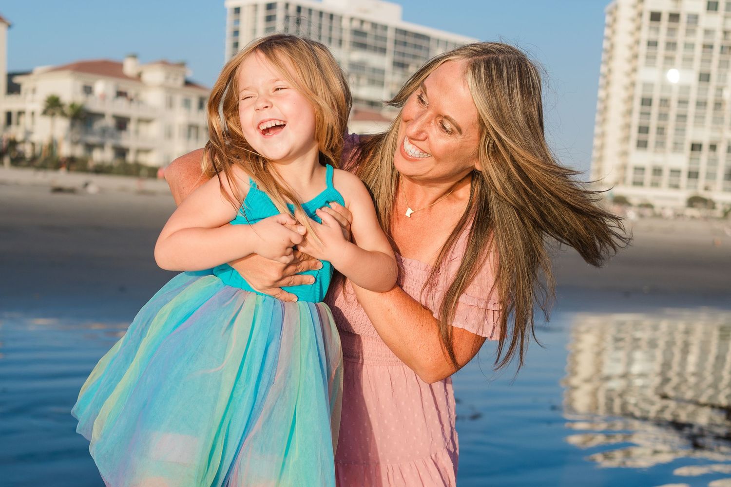 Two people share a playful moment laughing together on a beach with high-rise buildings in the background.