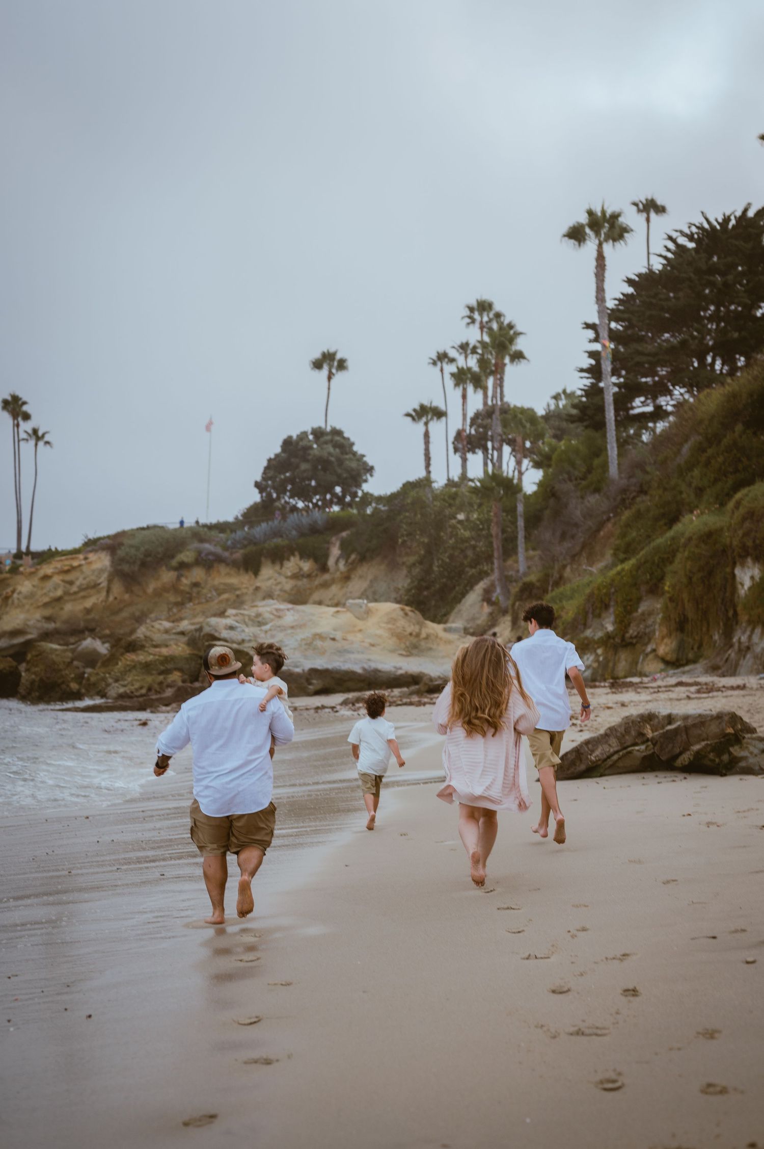 Group walking along beach with palm trees and cliffs in background during cloudy day.