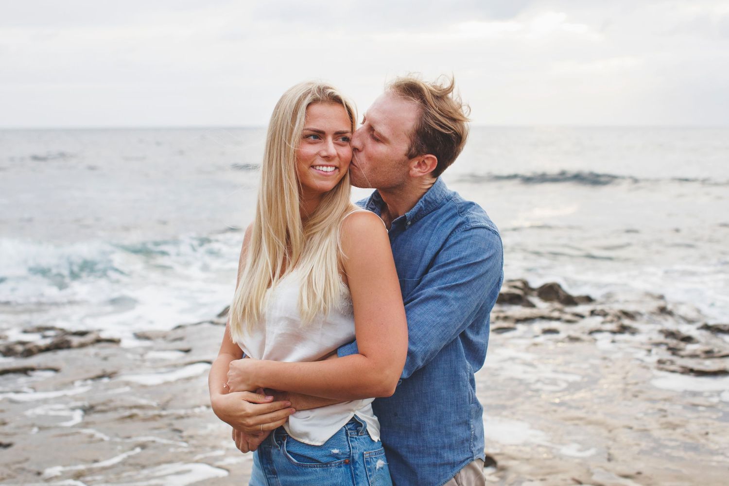 Intimate embrace on rocky shore with ocean waves in background.