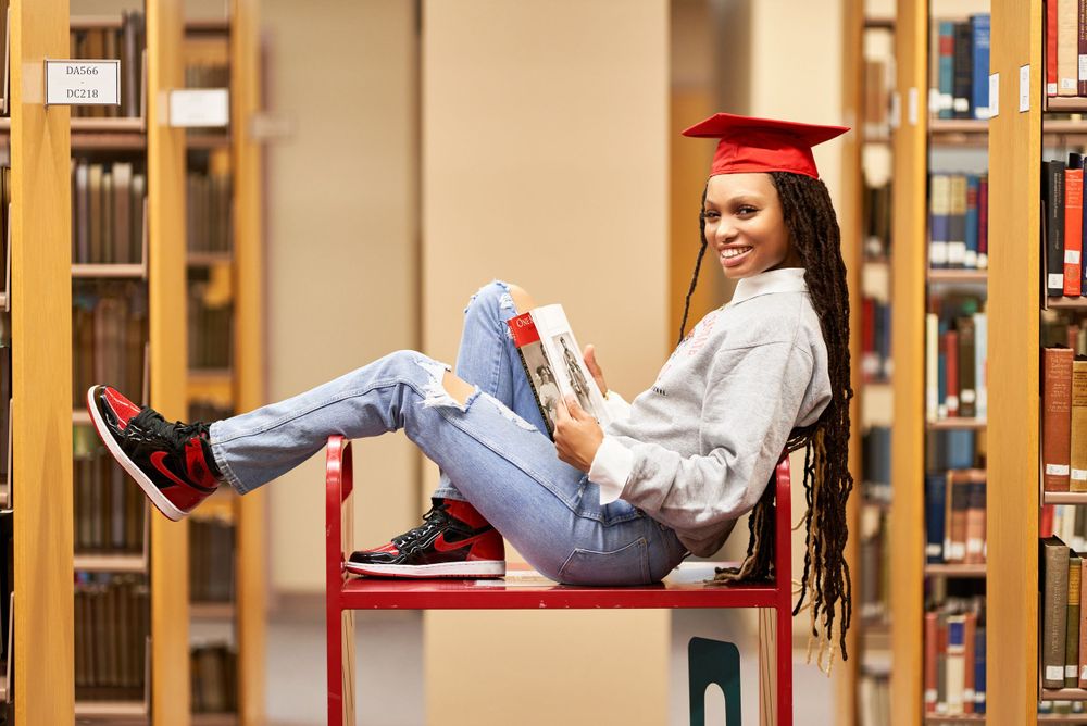 Graduate relaxing in library wearing red cap and gown while reading.