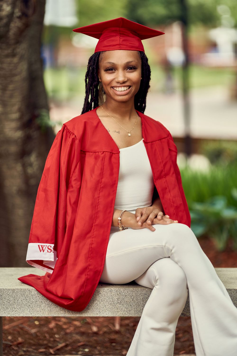Graduate posing in red cap and gown with white outfit sitting outdoors on campus.