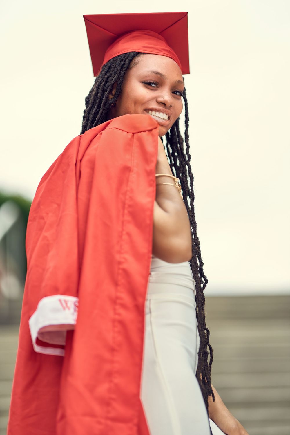 Graduate in red academic regalia poses with long braids against light background.