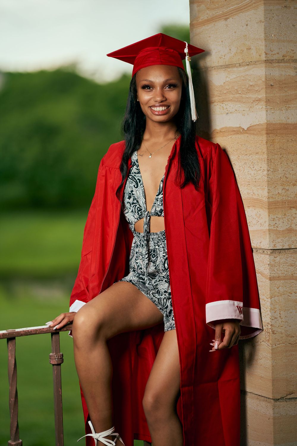 Graduate in red graduation gown posing by stone column.