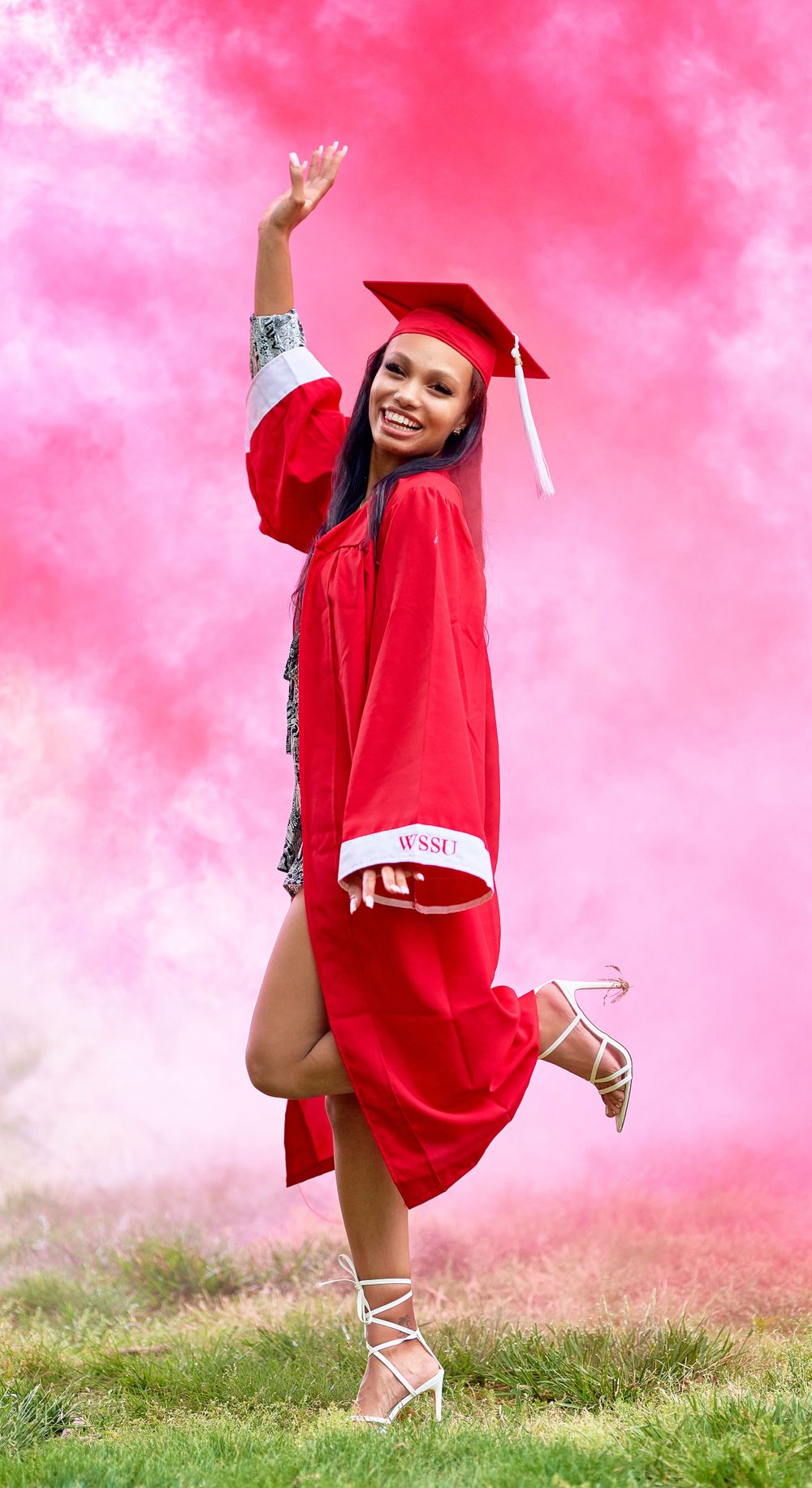 Graduate jumping joyfully in red cap and gown against pink sky background.