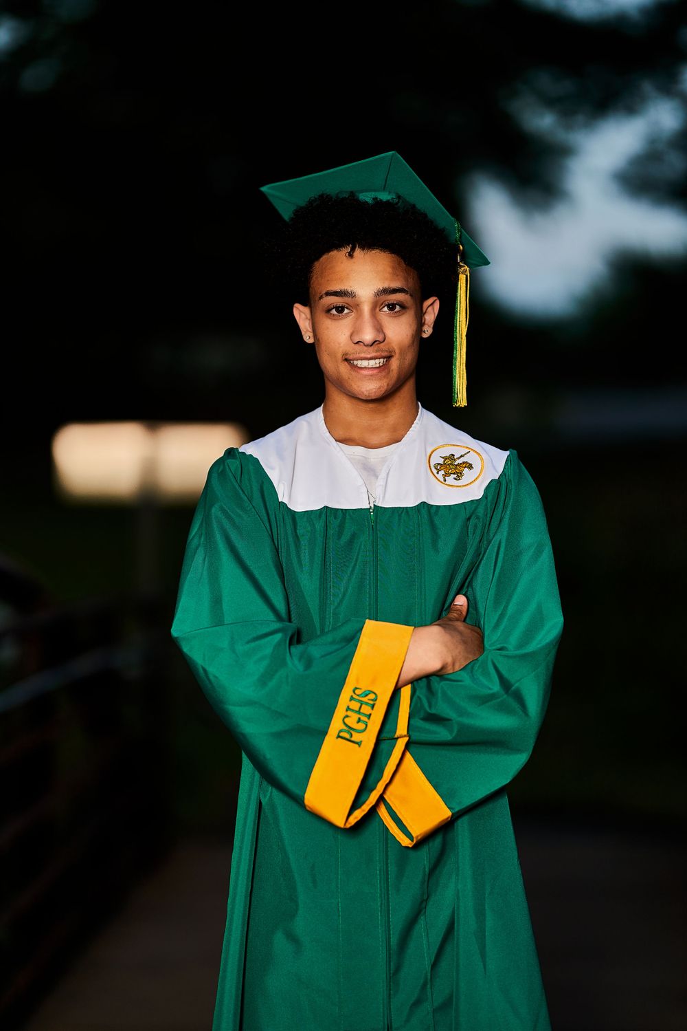 Graduate in dark green commencement gown standing outdoors at dusk.