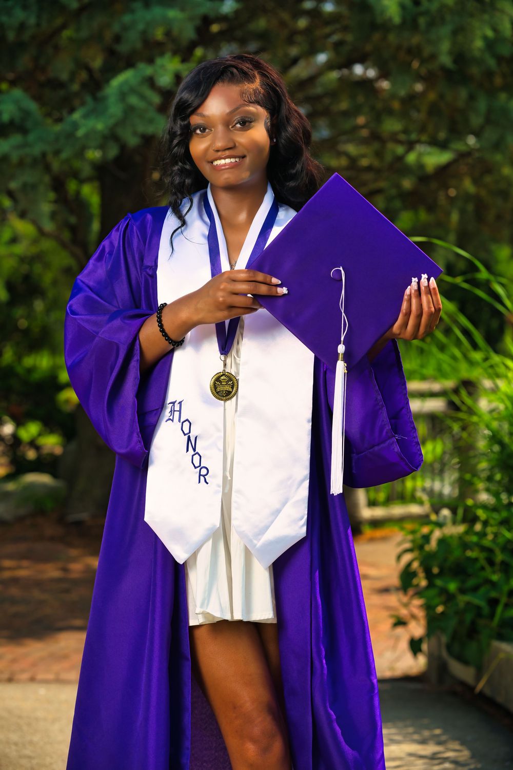 A graduate poses in a purple graduation gown and white stole against a green outdoor backdrop.
