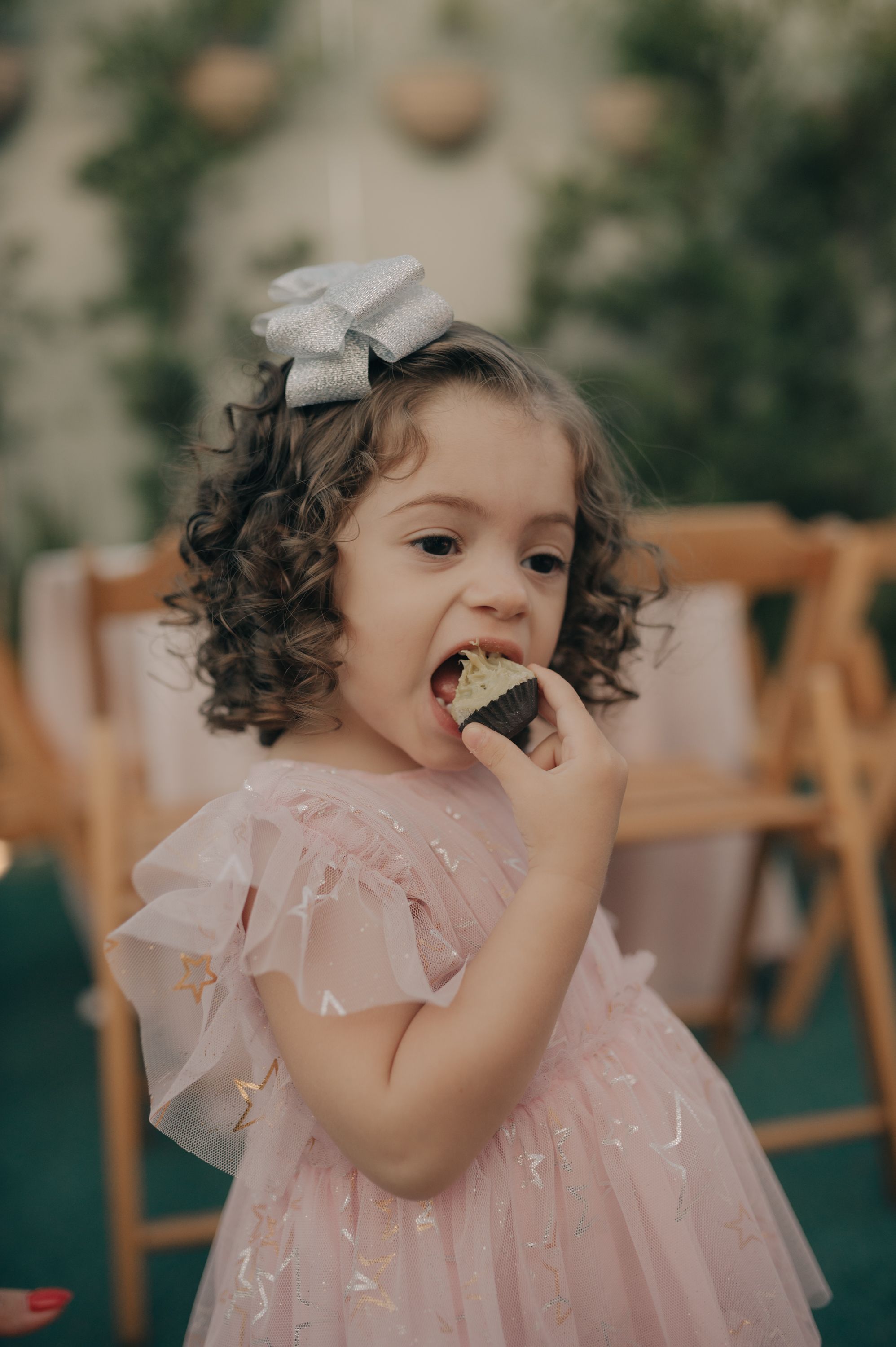 Young girl in pink dress eating dessert at party with curly hair and bow.