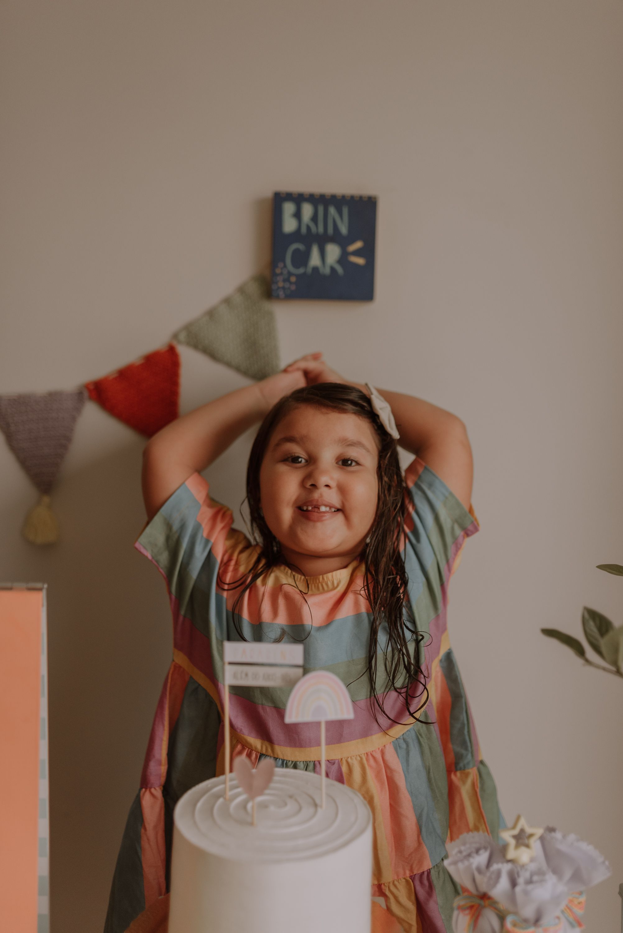 A child in a rainbow dress poses excitedly next to a white cake with a 'Rain Care' sign in the background.