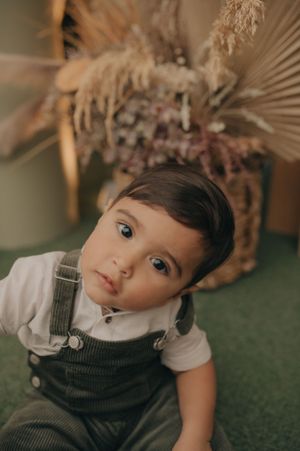 Young child in overalls sitting against dried floral arrangement background.