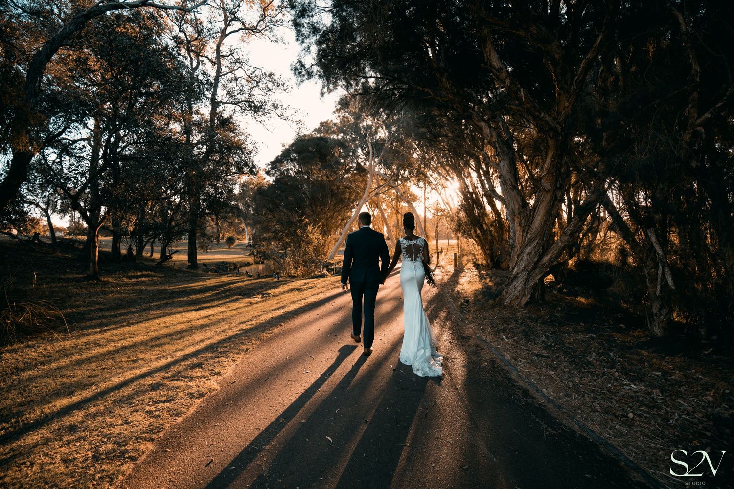 Silhouetted newlyweds walk hand in hand on a path during golden sunset.