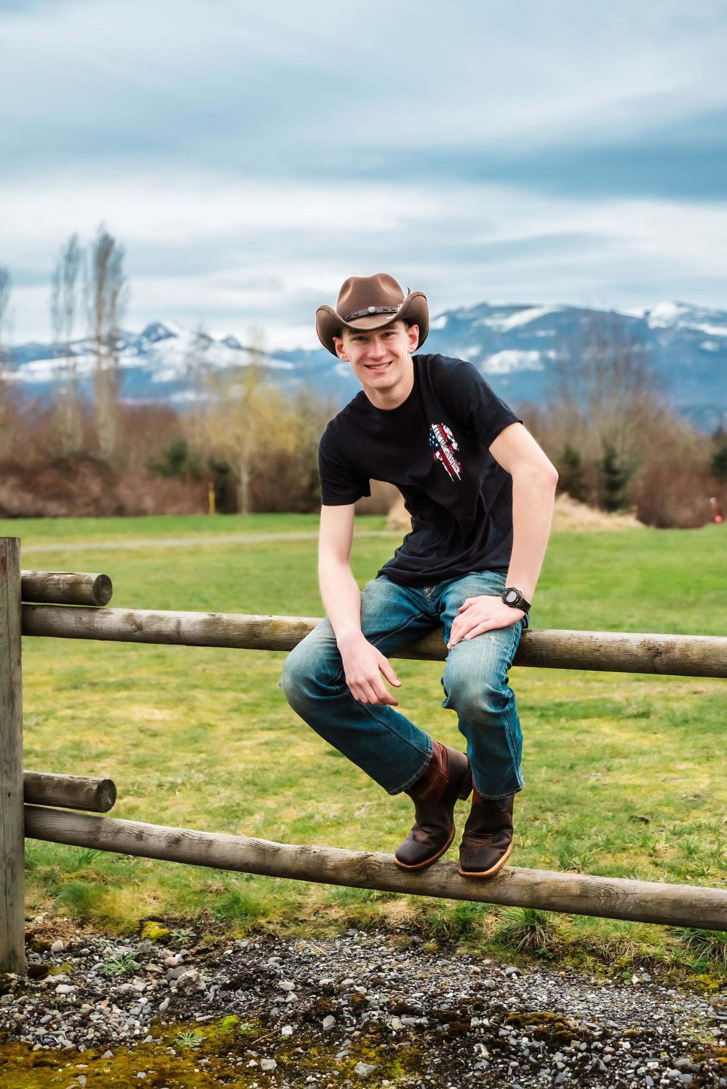 senior guy smiles sitting on fencepost backed by snow covered hills in skagit county