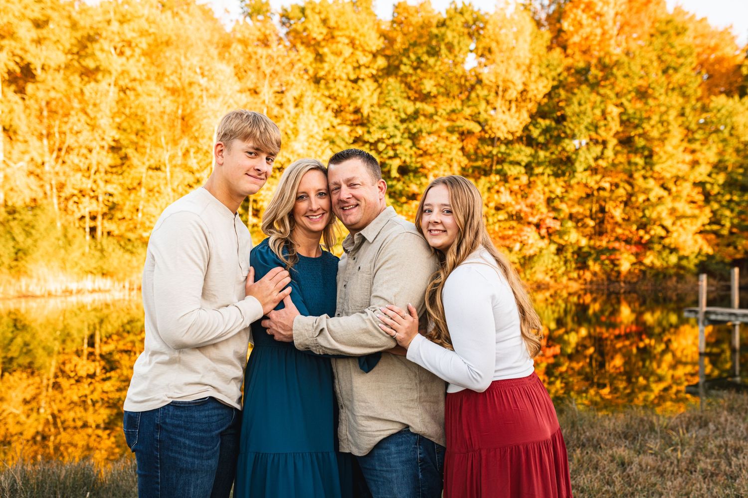 A family of 4 posing along a lake with the fall colors behind them
