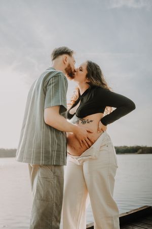 Couple kissing on a lakeside dock at sunset, woman with tattoos showing baby bump in white jeans.