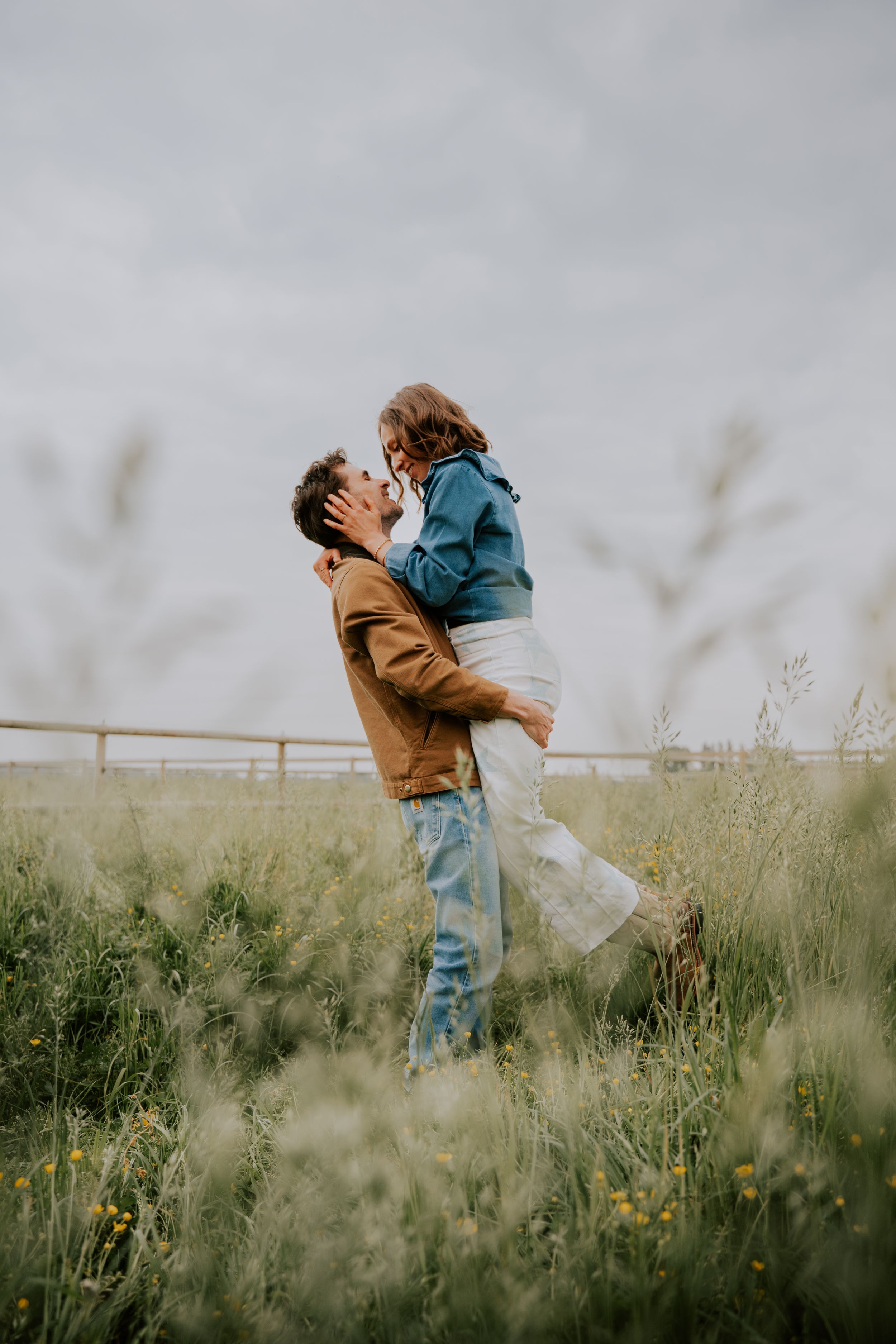 A man lifts a woman in a field of tall green grass, sharing a romantic moment under a cloudy sky.