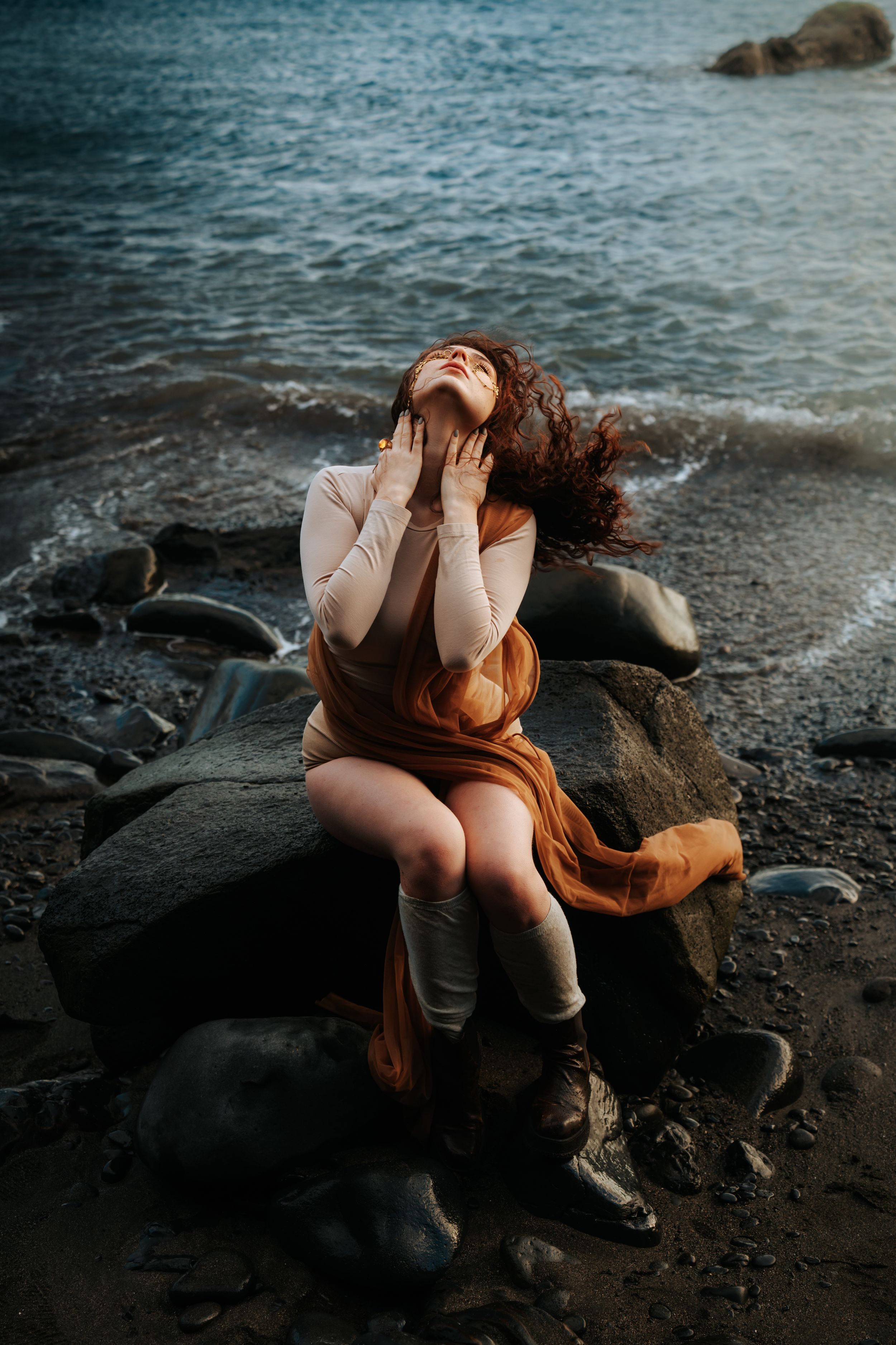A woman with flowing red hair sits on coastal rocks, hands on face, wearing a rust skirt and white top by the ocean.