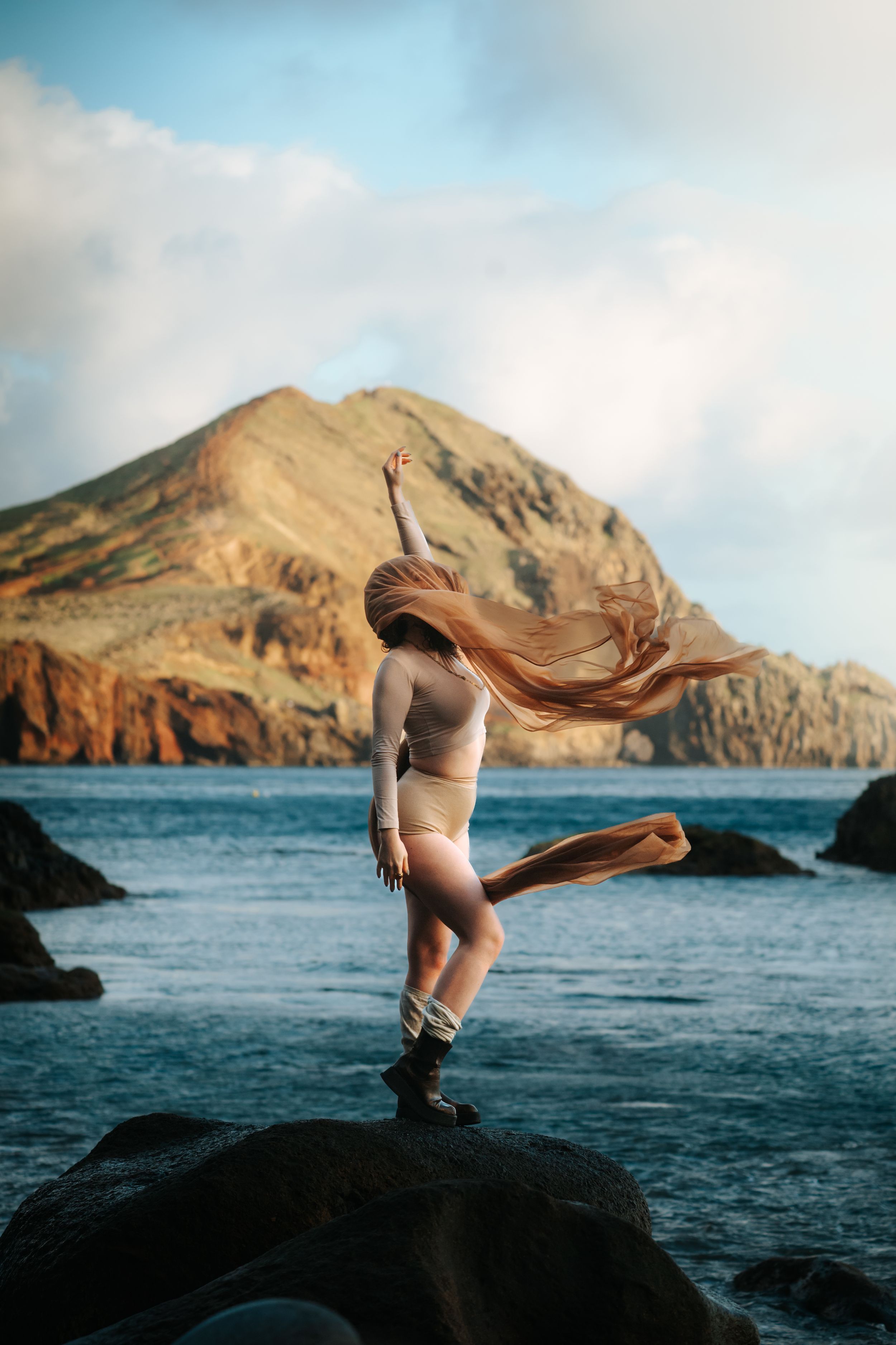 Woman dancing on coastal rocks with hair flowing in wind, dramatic mountain and ocean backdrop.