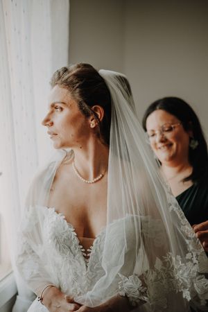 Bride in lace wedding gown and veil gazes out window as smiling bridesmaid adjusts her veil behind her.
