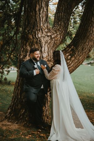Bride and groom share a romantic moment leaning against a large tree in a lush green park on their wedding day.