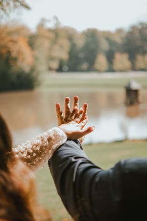 Couple holding hands outdoors near a lake in autumn, showcasing wedding rings on their fingers.
