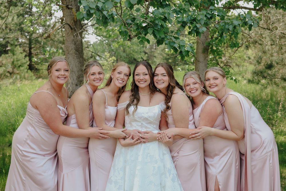 Wedding party in light pink bridesmaid dresses poses together in a lush outdoor garden setting.