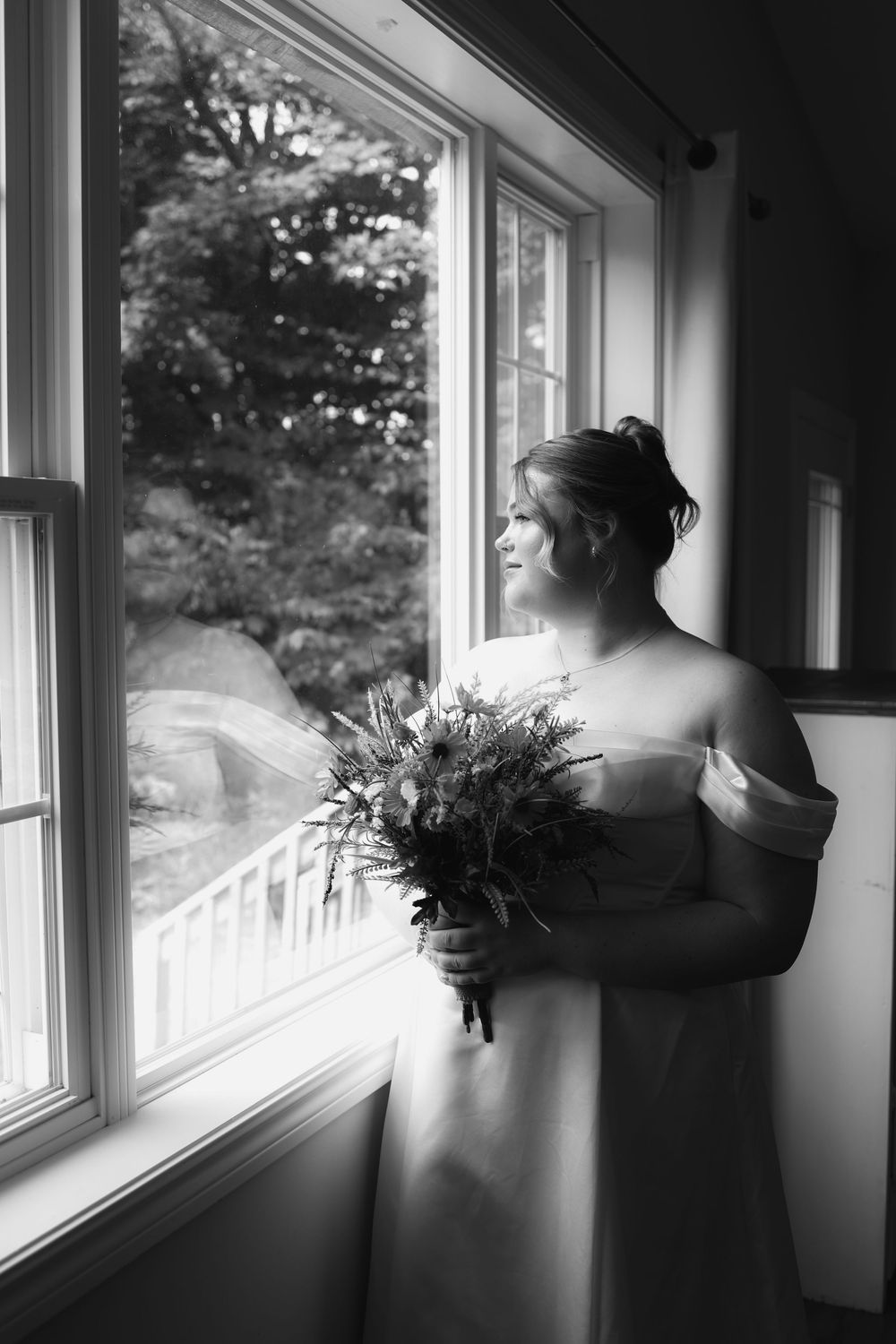 A person in a flowing white gown gazes thoughtfully out a window while holding a bouquet of flowers.