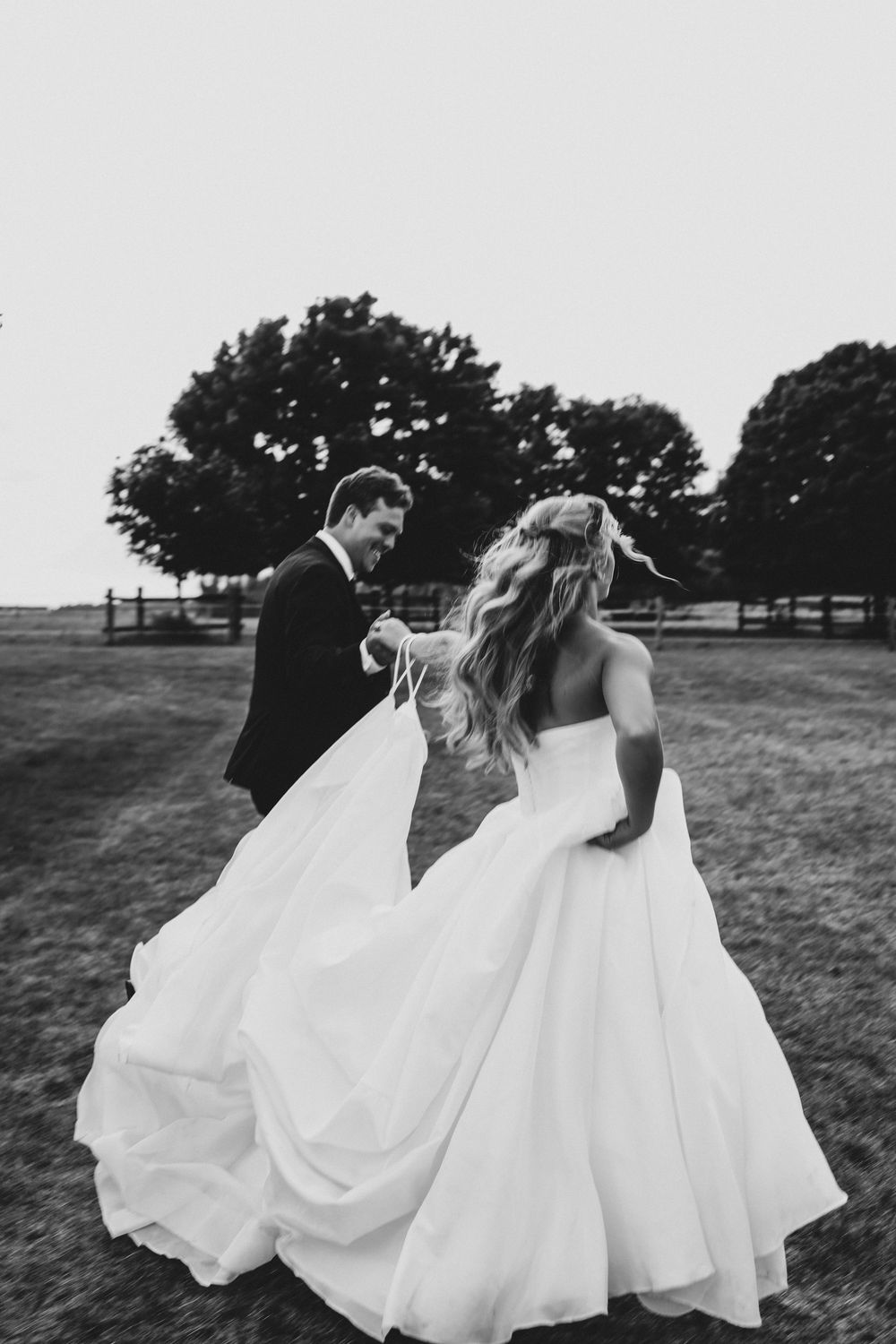A couple in wedding attire dances and spins joyfully in a grassy field with trees in the background.