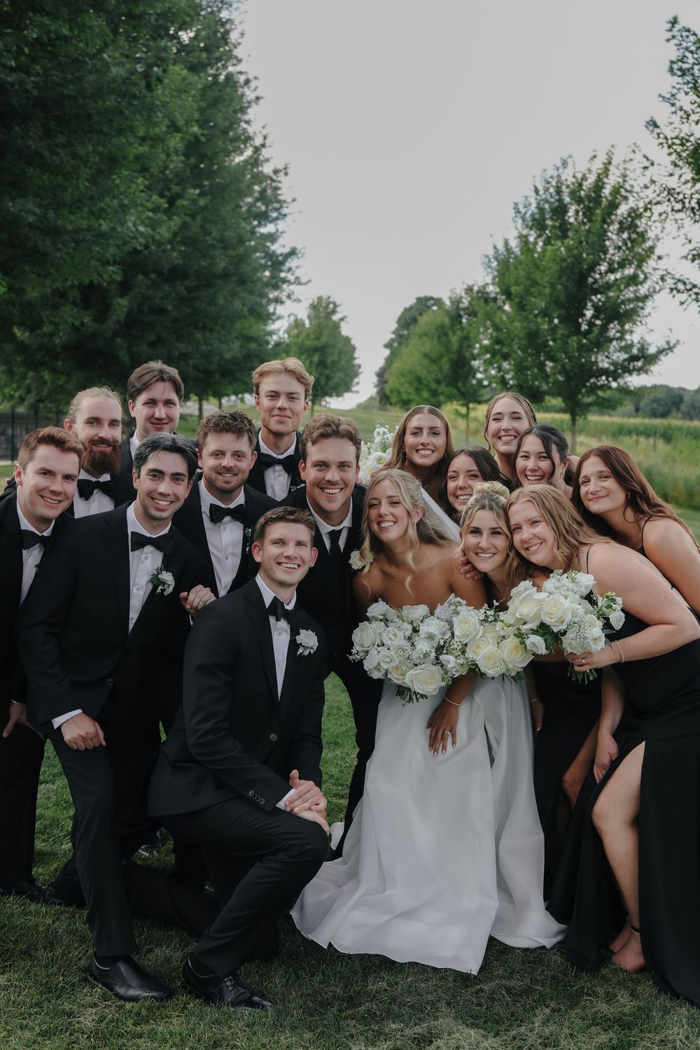 Wedding party in black attire and formal dresses poses together on grass with trees in background.