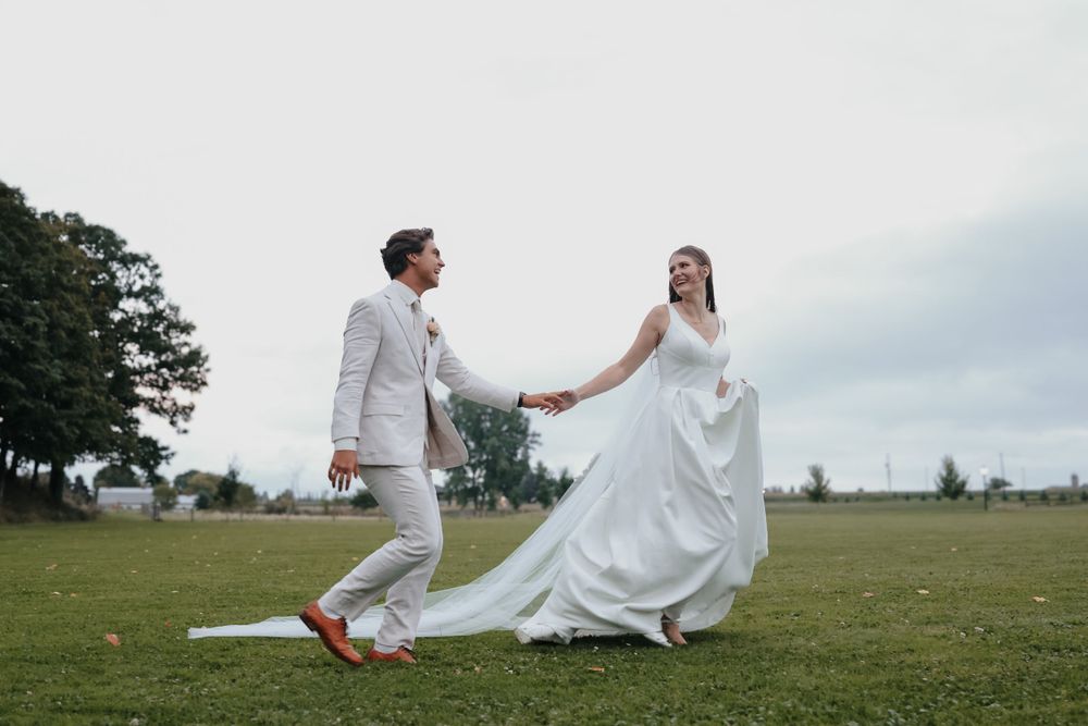 A newlywed couple dances playfully on a grassy field in white wedding attire.