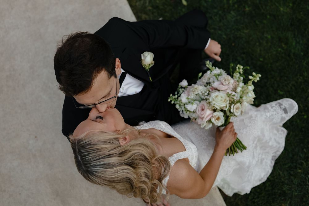 Aerial view of newlyweds sharing a romantic moment while holding a delicate pink and white floral bouquet.
