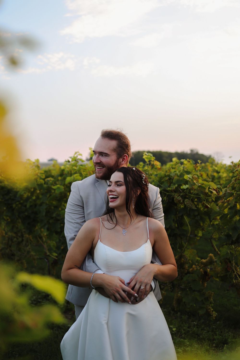 A newlywed couple embraces and laughs together in a vineyard at sunset.