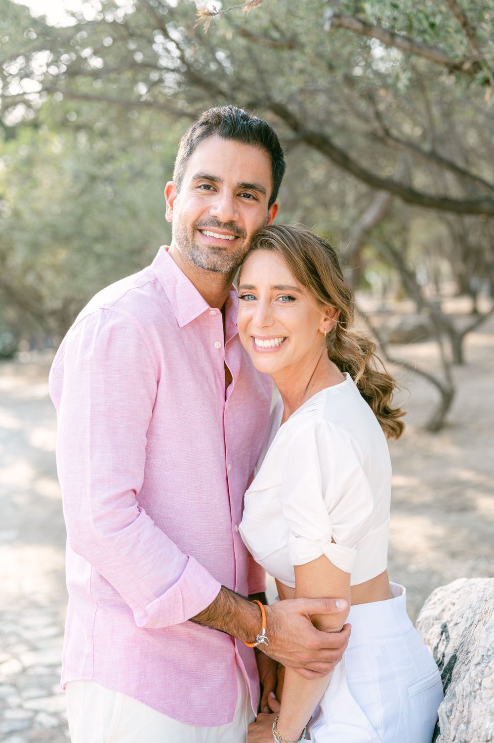 A couple smiles while embracing outdoors wearing pink and white casual attire.
