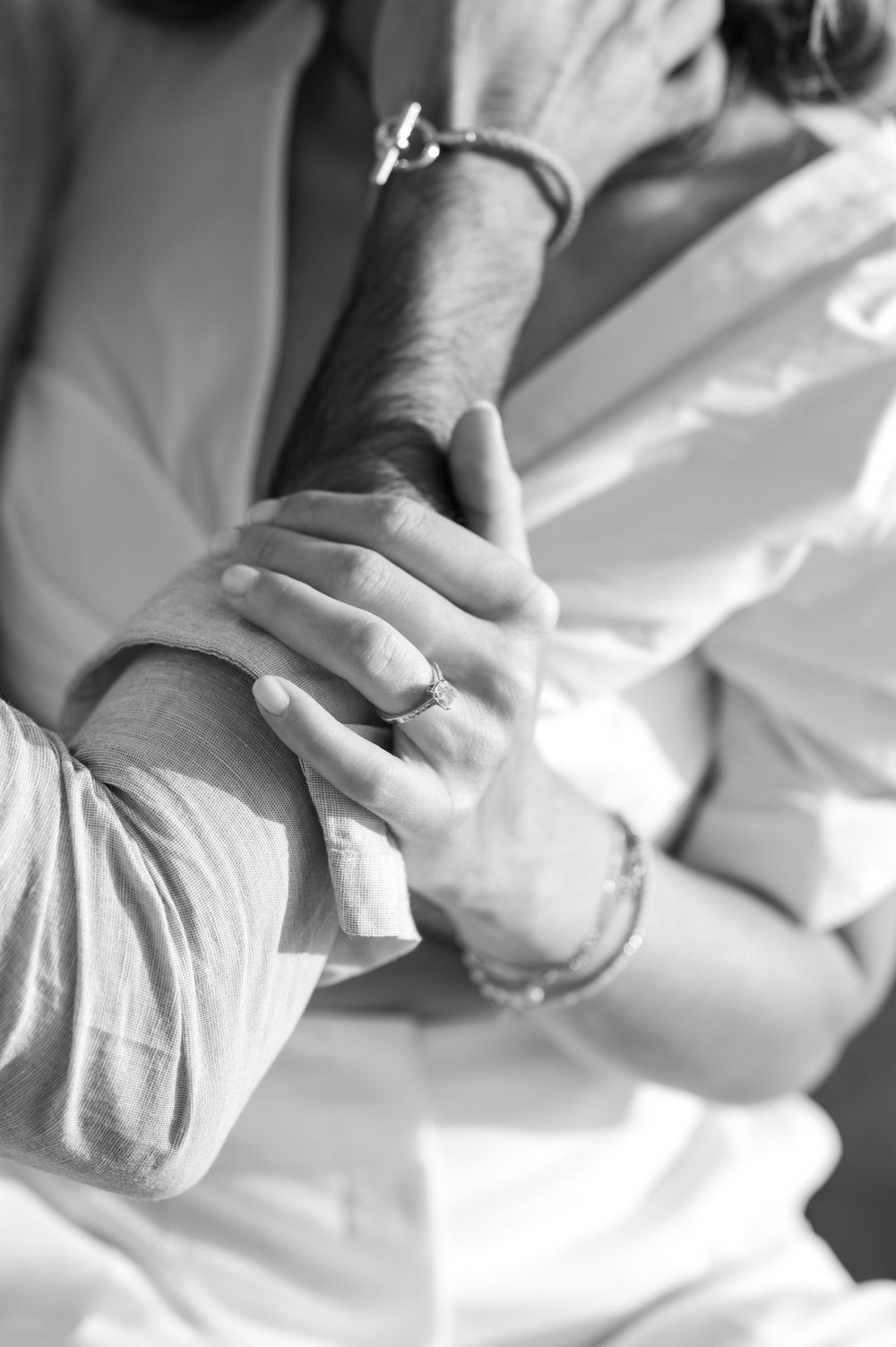Close-up black and white photo of hands clasped together showing engagement ring and bracelet details.