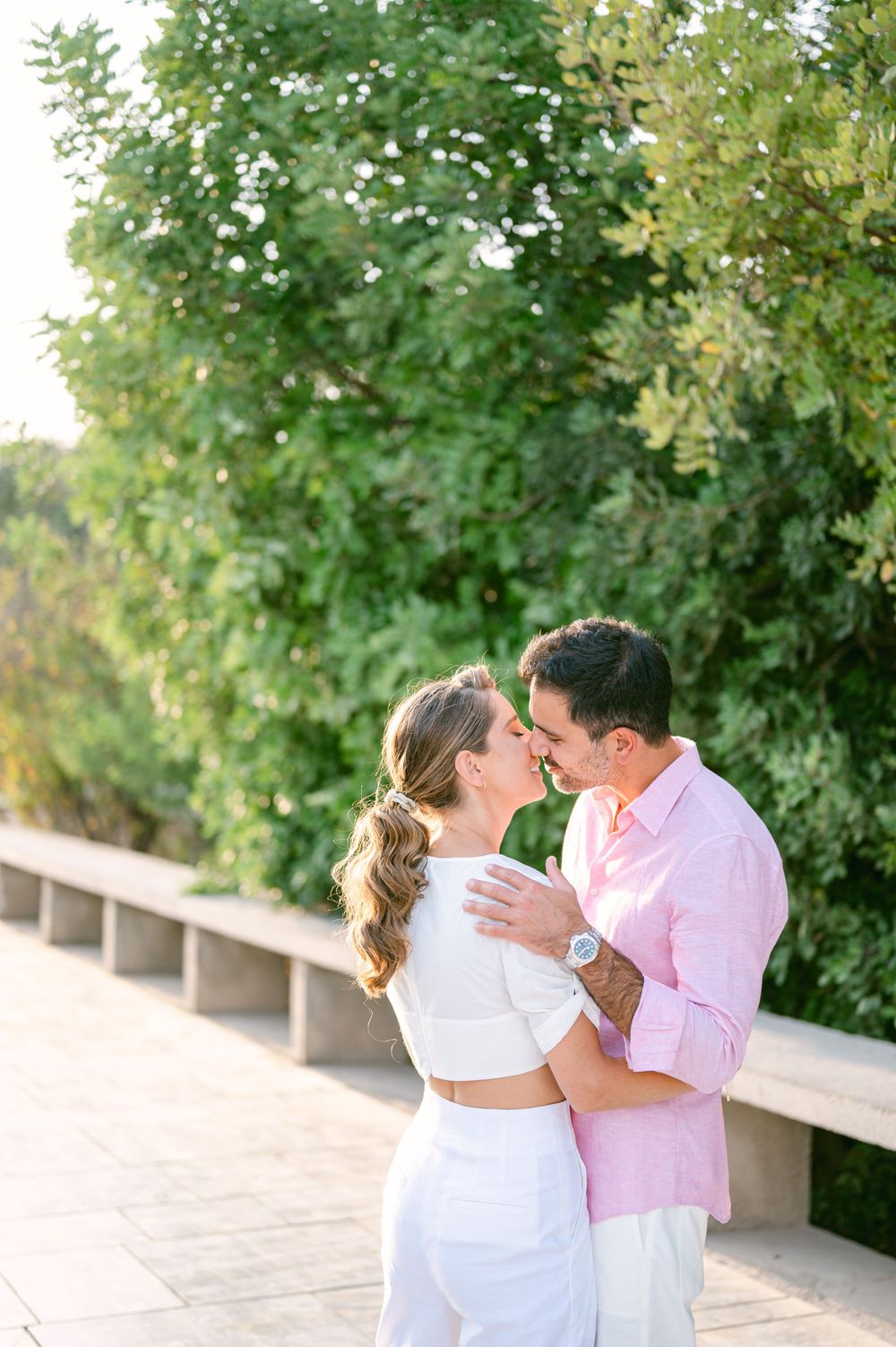 Romantic couple embracing in white and pink outfits against lush green garden backdrop.