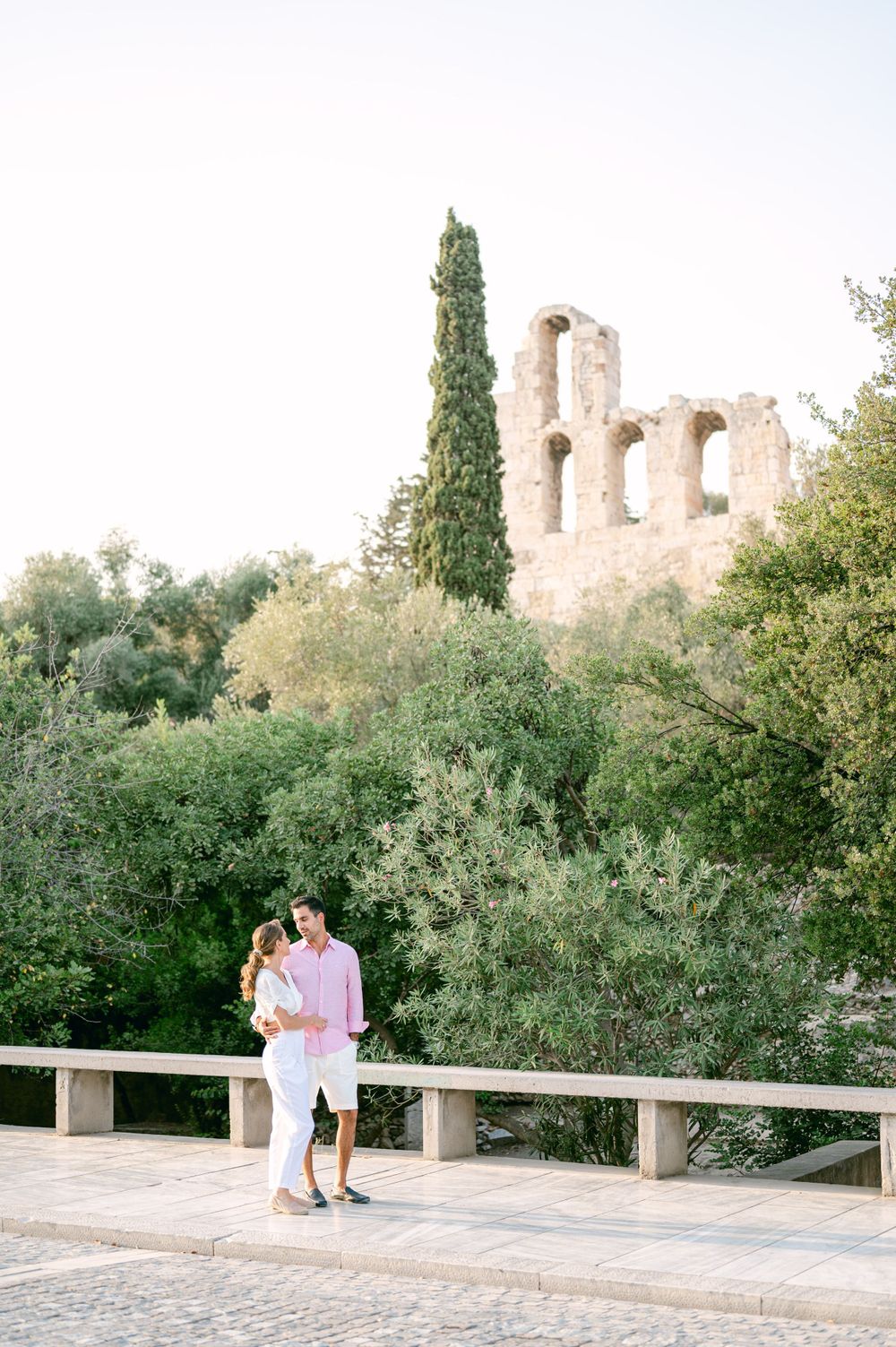 Couple sharing romantic moment near ancient ruins with cypress trees in Mediterranean setting.