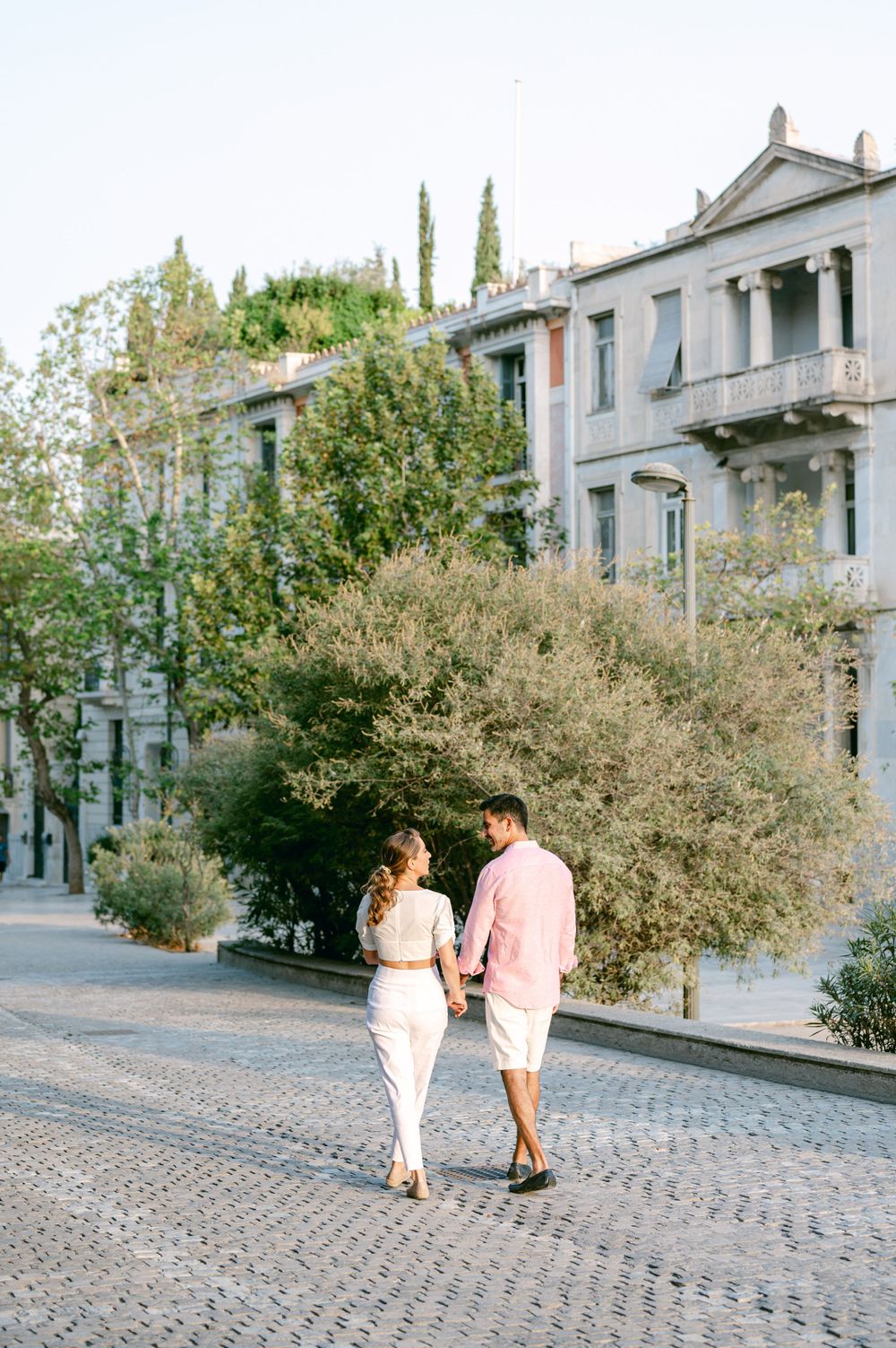 Couple walking hand in hand along cobblestone street with classic European architecture.