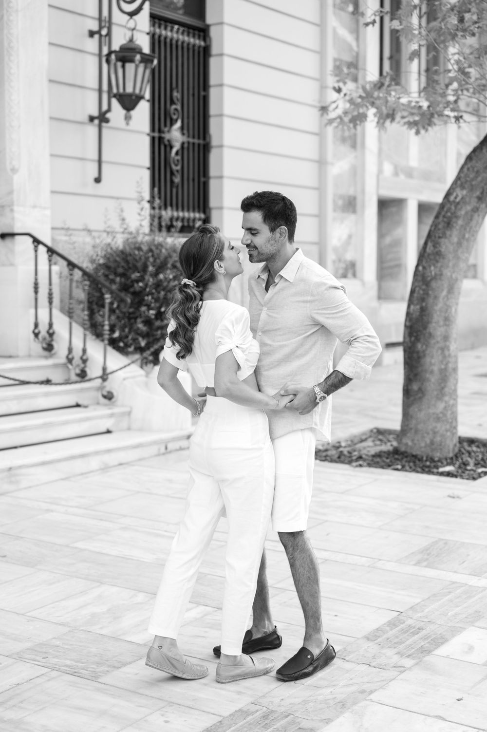 Black and white photo of couple dancing on elegant urban sidewalk in matching white outfits.