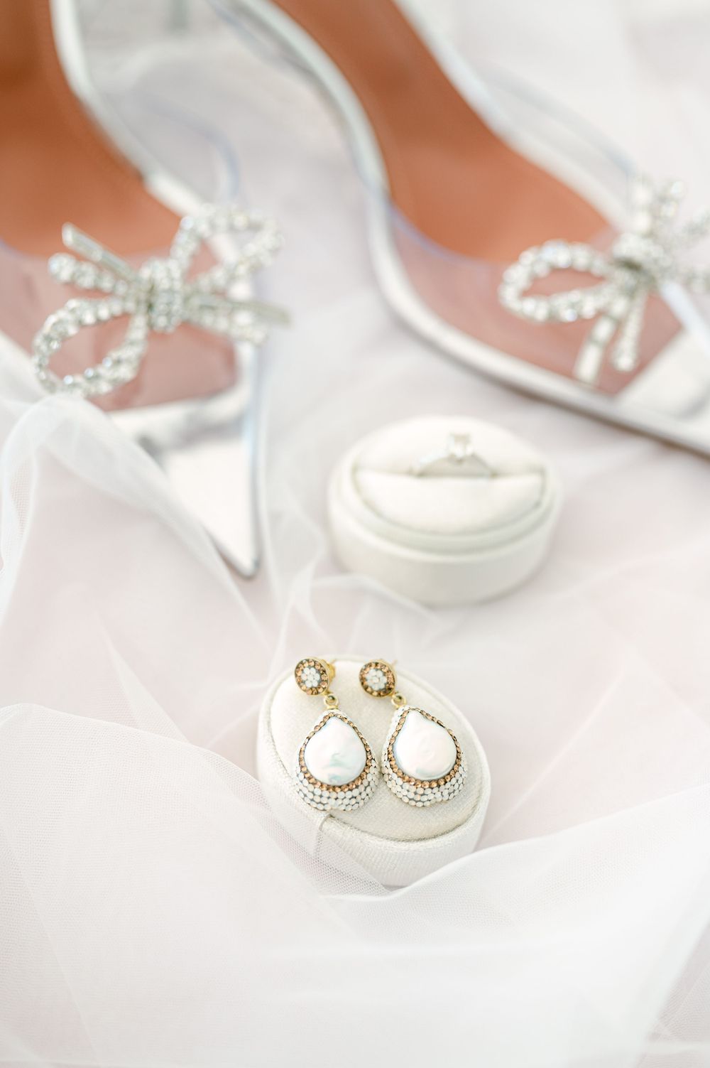 Elegant bridal accessories including crystal bow heels and pearl drop earrings on white fabric.