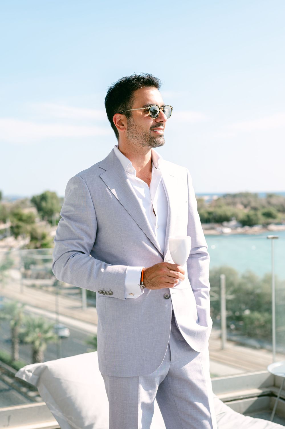 A sophisticated figure in a light grey suit and sunglasses poses on a balcony overlooking a coastal landscape.