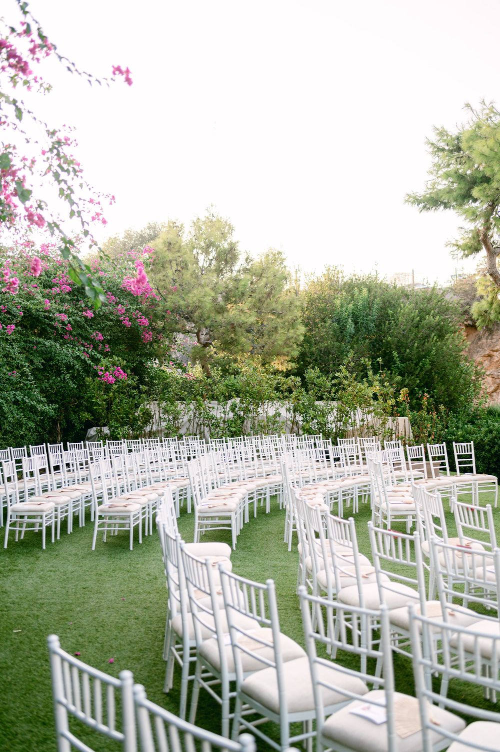 Rows of white chiavari chairs set up for an outdoor wedding ceremony with flowering bushes in the background.