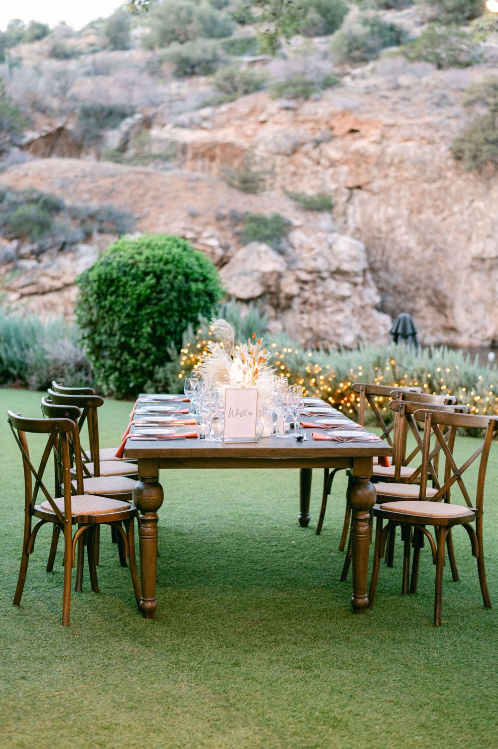 A romantic dining table setup on a lawn with wooden chairs and candles against a rocky backdrop.