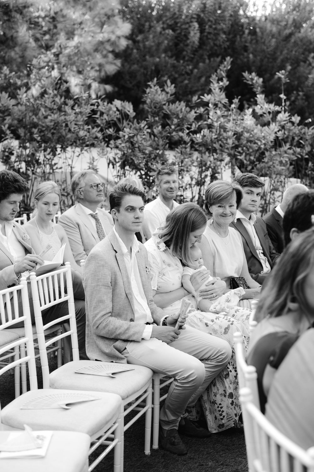 Wedding guests seated in white chairs during an outdoor ceremony captured in black and white photography.
