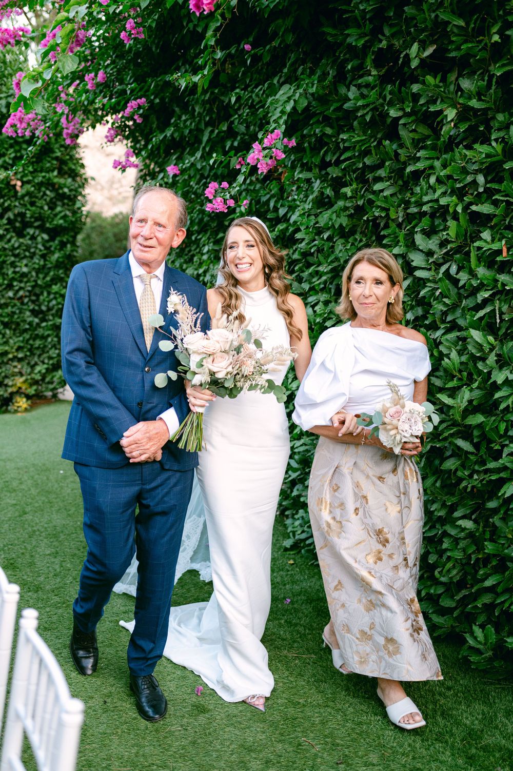 A wedding processional in a garden setting with flowering pink bushes and white chairs lining the path.