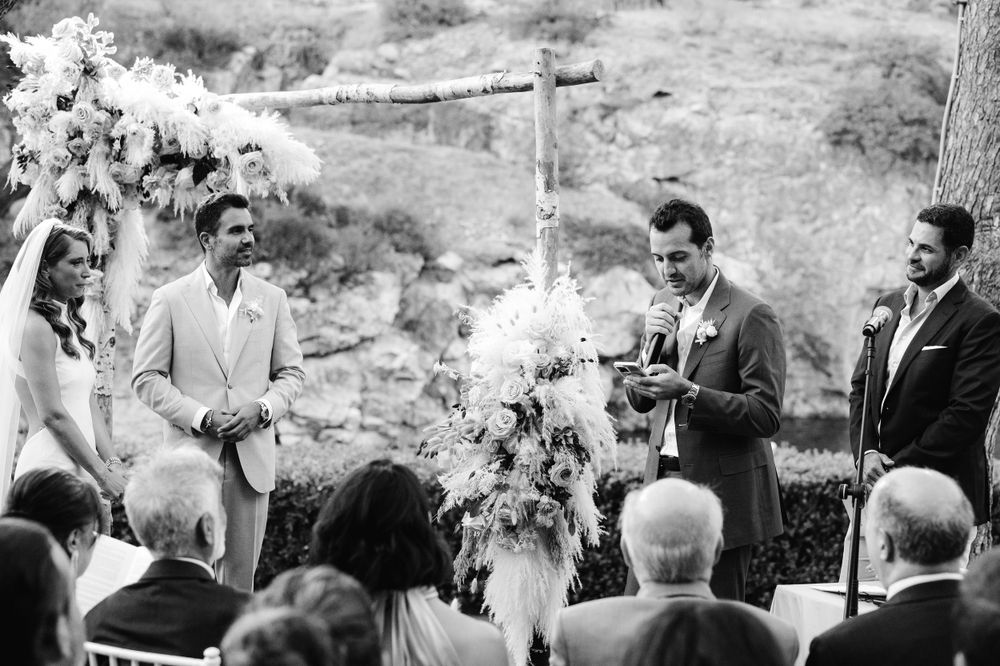 An outdoor wedding ceremony takes place under a floral arch with guests seated on a hillside.