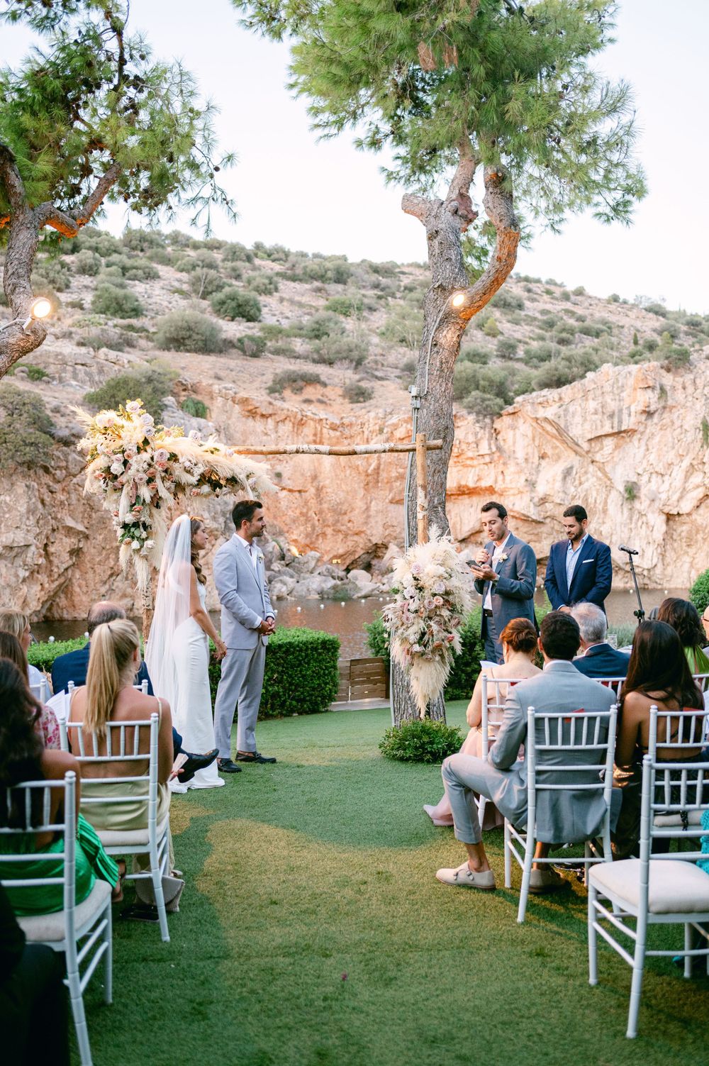 A scenic outdoor wedding ceremony with white chairs and floral arrangements against desert cliffs.