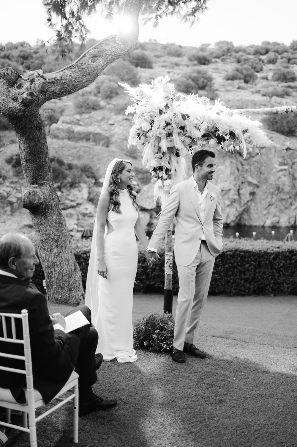 A black and white photo captures an intimate outdoor wedding ceremony moment beneath a tree.
