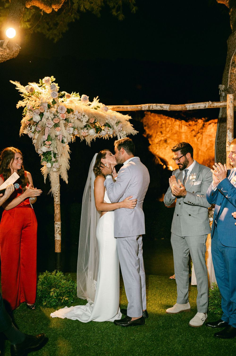 A nighttime wedding ceremony under a beautifully decorated floral arch with guests watching.