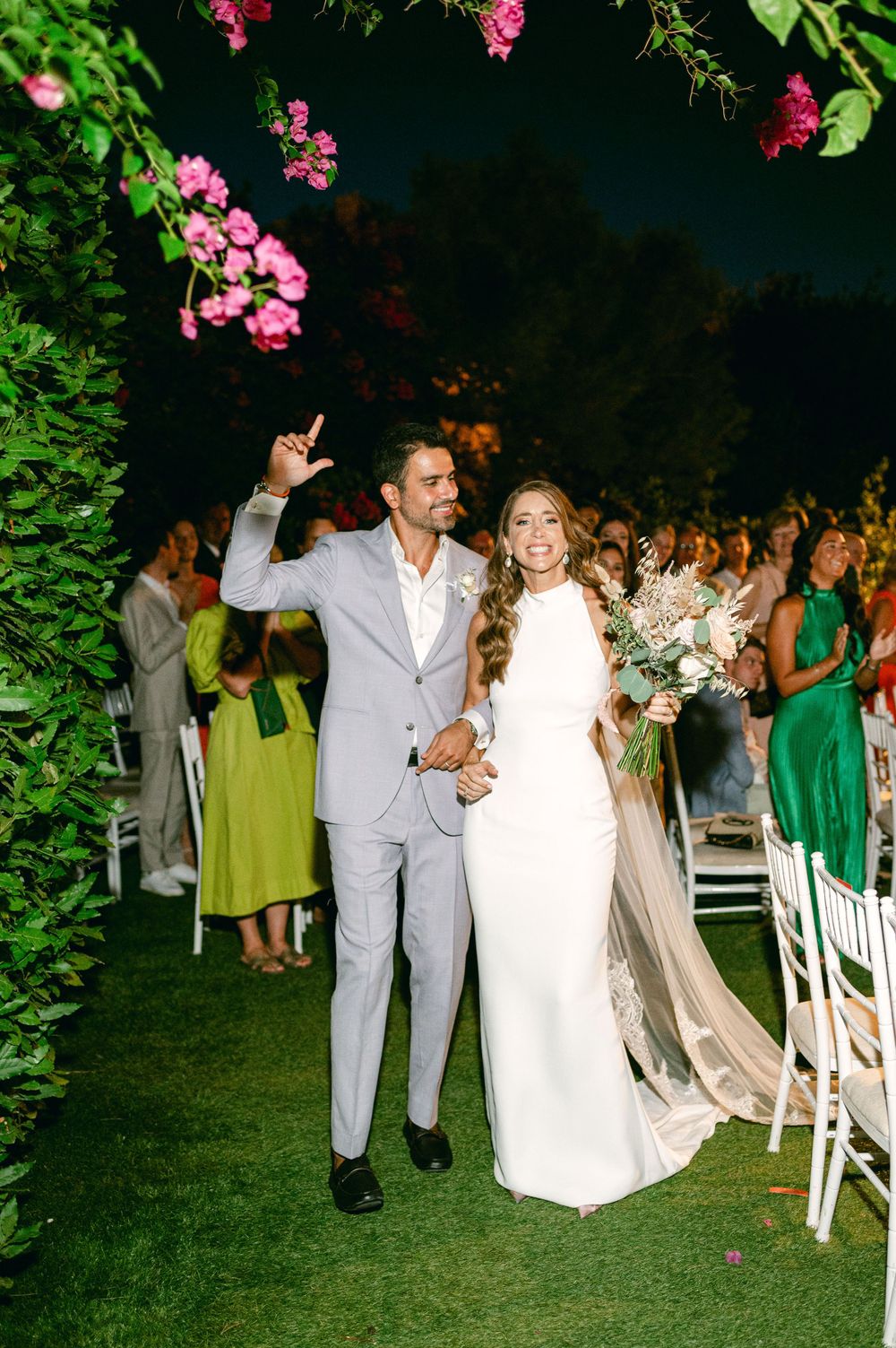 A joyful couple walks down the aisle while guests in colorful dresses look on.