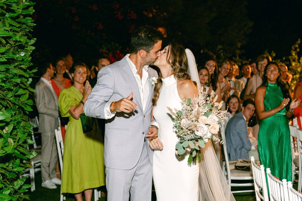 A celebratory moment between newlyweds as wedding guests in green and yellow dresses look on.
