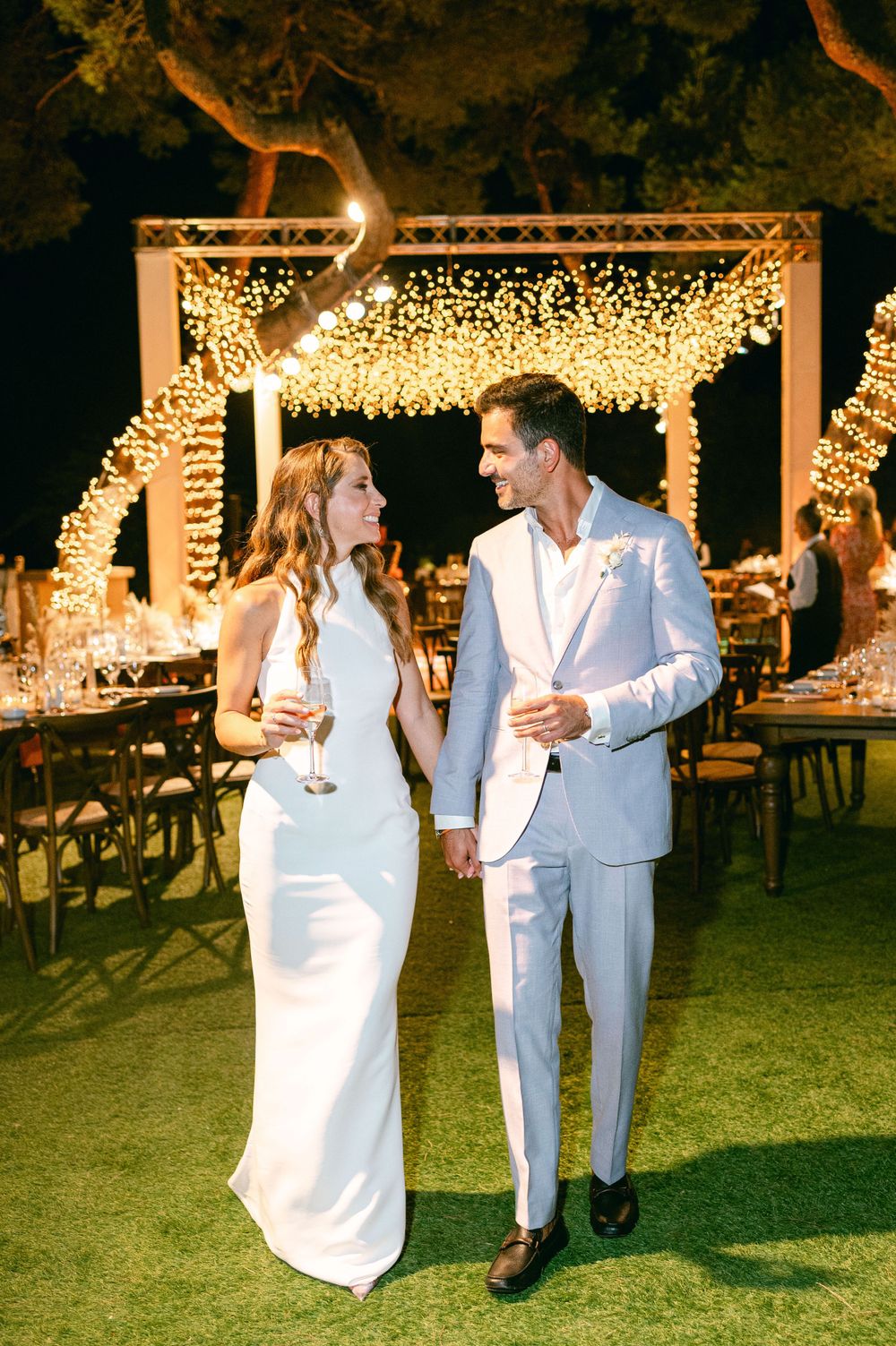 Elegant wedding couple in matching white attire share a moment under a twinkling light canopy at night.