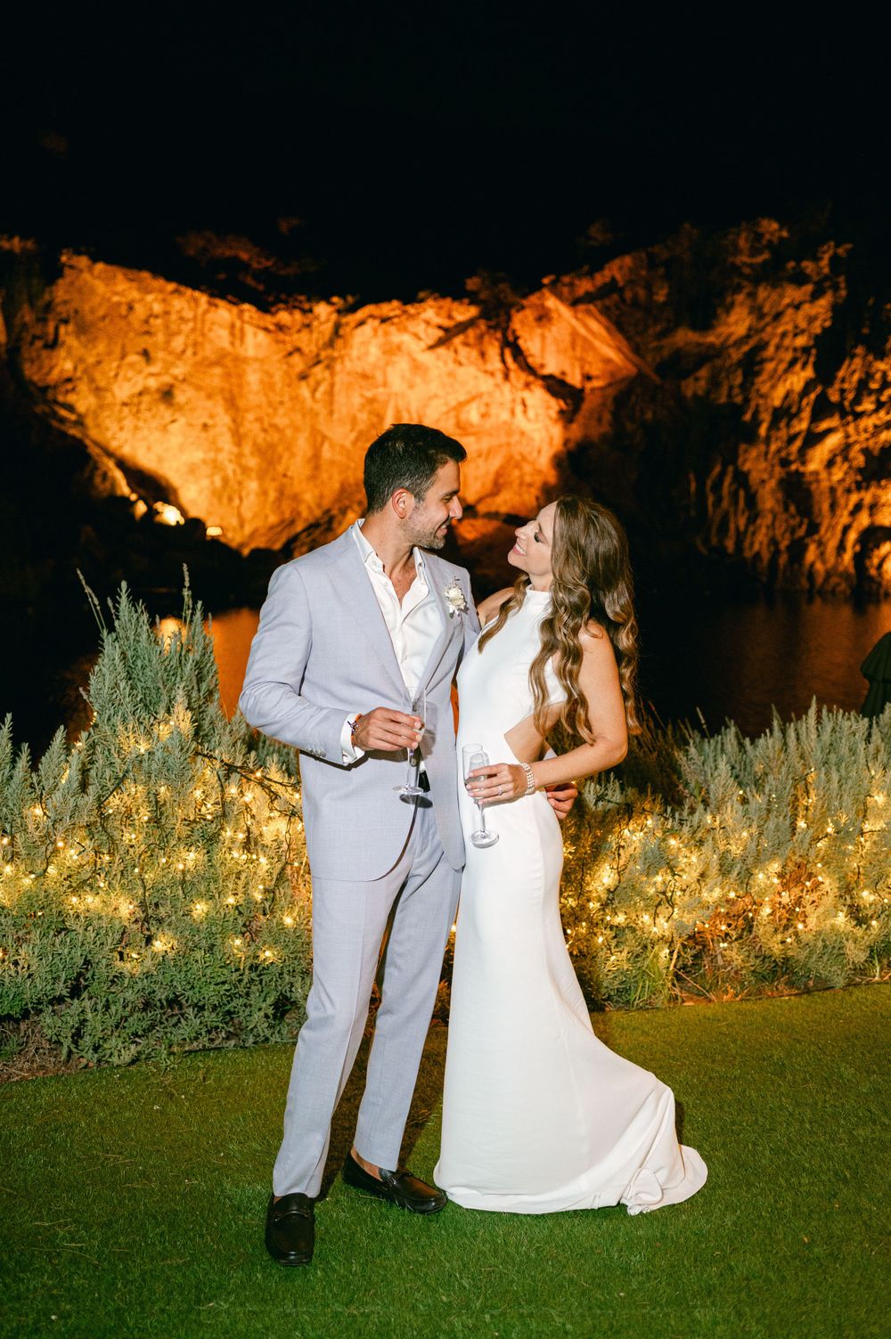 Romantic wedding scene with couple in white outfits against illuminated cliff face at night.